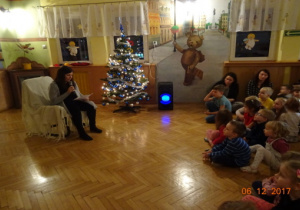 Choinka, mama przedszkolaka siedzi na fotelu i czyta dzieciom bajkę. Dzieci słuchają czytanego tekstu.